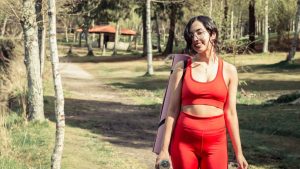 Smiling woman in red sport clothing holding a yoga mat outdoors.